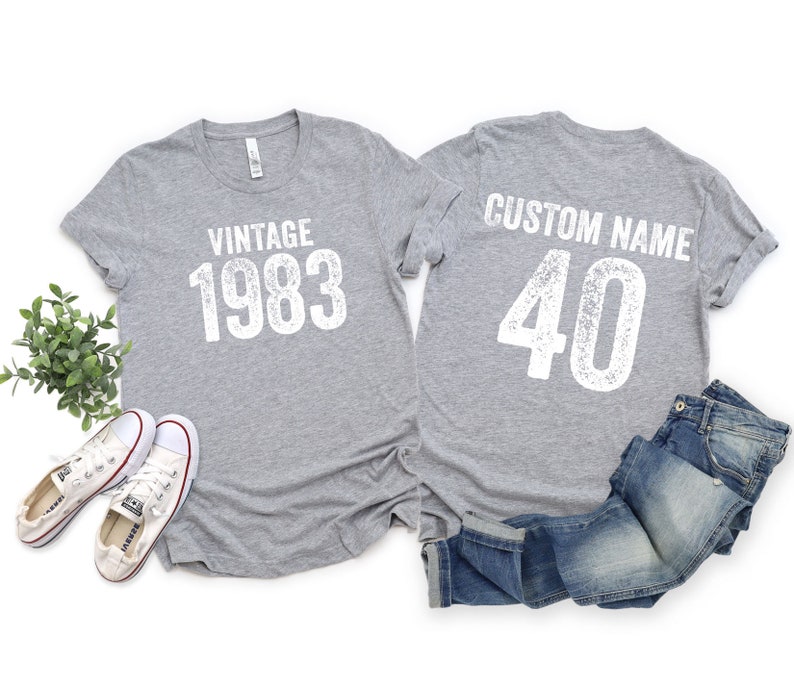 K&ouml;nnte beinhalten: Zwei graue T-Shirts mit wei&szlig;em Text. Das vordere Shirt sagt "Vintage 1983" und das hintere Shirt sagt "Custom Name 40".