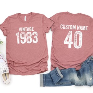 K&ouml;nnte beinhalten: Zwei heidefarbene T-Shirts mit wei&szlig;em Text. Das vordere Shirt sagt "Vintage 1983". Das hintere Shirt sagt "Benutzerdefinierter Name 40".