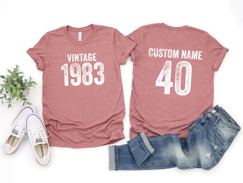 K&ouml;nnte beinhalten: Zwei heidefarbene T-Shirts mit wei&szlig;em Text. Das vordere Shirt sagt "Vintage 1983". Das hintere Shirt sagt "Benutzerdefinierter Name 40".