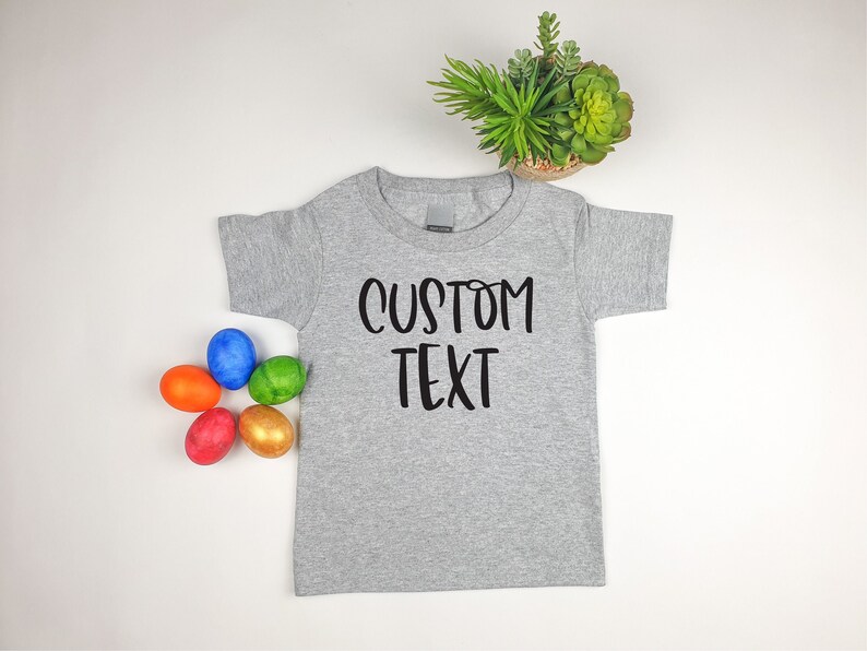 Puede incluir: Una camiseta gris de manga corta con el texto "CUSTOM TEXT" impreso en negro. La camiseta est&aacute; colocada sobre una superficie blanca con cinco huevos de Pascua de colores.