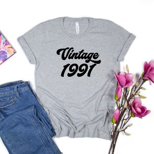 May include: Gray t-shirt with black text that reads "Vintage 1991" in a retro font. The shirt is folded and laid flat on a white background with a pair of blue jeans and a floral planner to the left and a sprig of pink flowers to the right.
