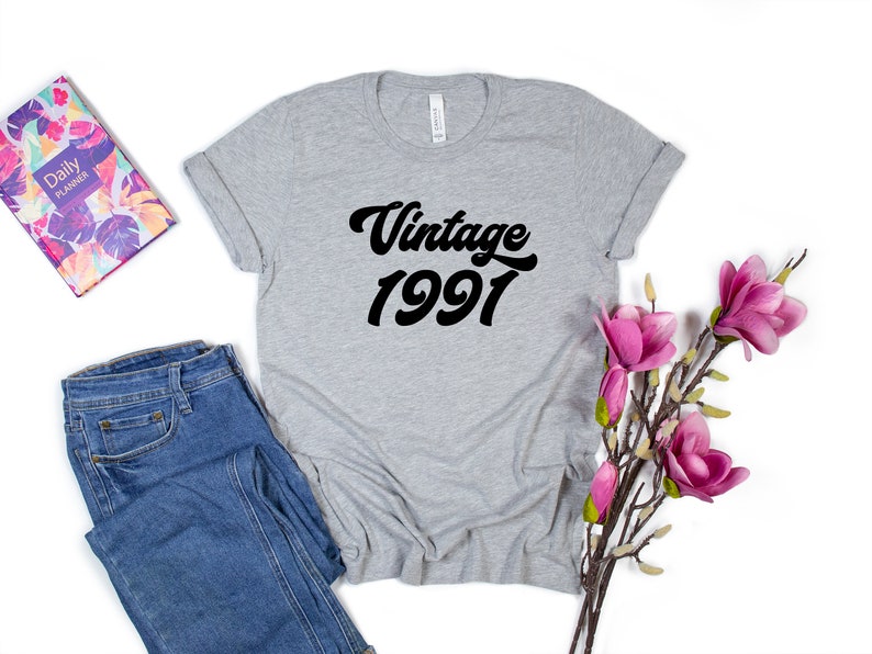 May include: Gray t-shirt with black text that reads "Vintage 1991" in a retro font. The shirt is folded and laid flat on a white background with a pair of blue jeans and a floral planner to the left and a sprig of pink flowers to the right.