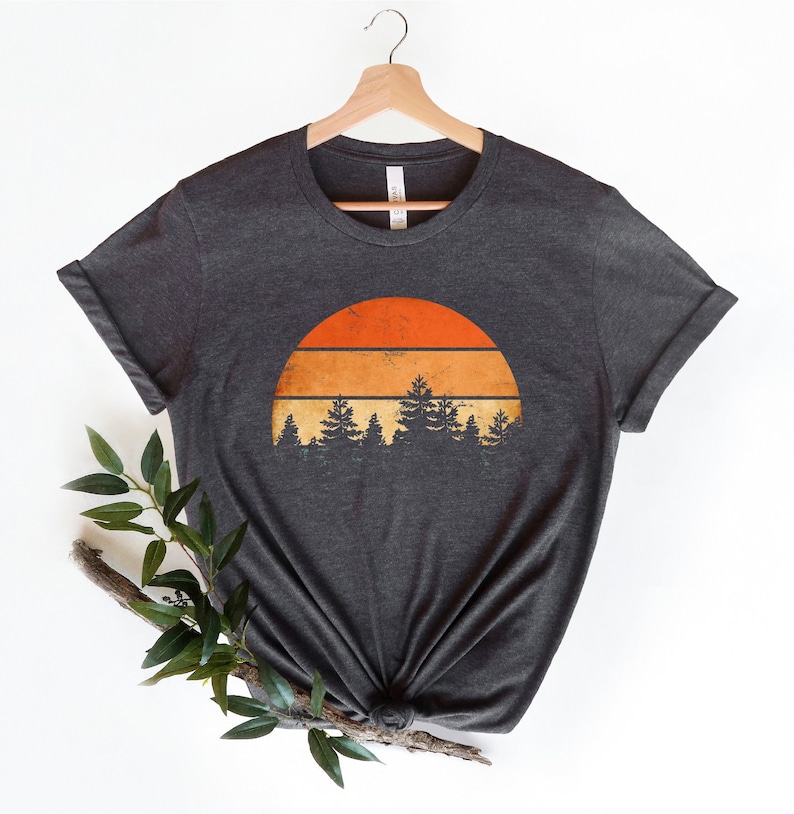 May include: A dark heather grey t-shirt featuring a vintage sunset graphic with a half-circle orange sun with three horizontal stripes and a row of dark green pine trees in front of it.
