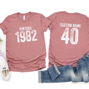 May include: Two t-shirts, one with the text "Vintage 1982" and the other with the text "Custom Name 40". The shirts are a dusty rose color and are laid out on a white surface with a pair of white sneakers and a pair of blue jeans.