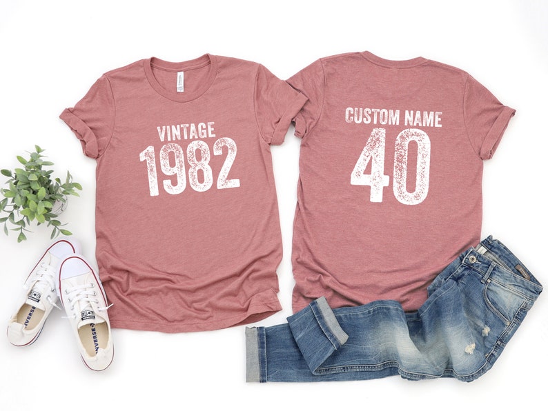 May include: Two t-shirts, one with the text "Vintage 1982" and the other with the text "Custom Name 40". The shirts are a dusty rose color and are laid out on a white surface with a pair of white sneakers and a pair of blue jeans.