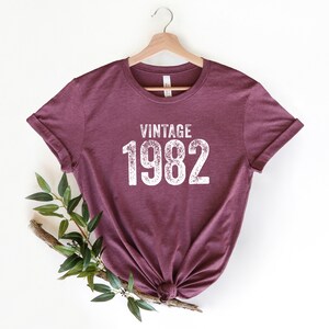 May include: A maroon t-shirt with a white vintage 1982 graphic.