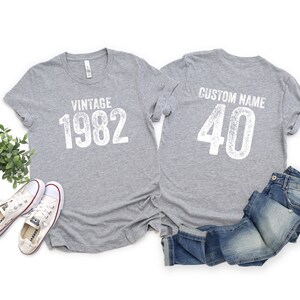 May include: Two gray t-shirts with white text. The front shirt says "Vintage 1982" and the back shirt says "Custom Name 40".
