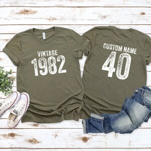 May include: Two olive green t-shirts with white text. One shirt says "Vintage 1982" and the other says "Custom Name 40".