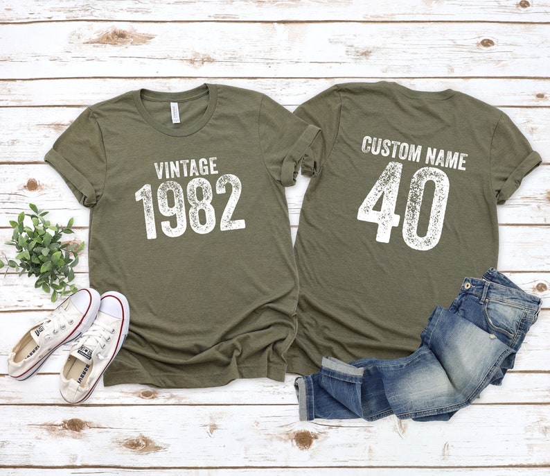 May include: Two olive green t-shirts with white text. One shirt says "Vintage 1982" and the other says "Custom Name 40".