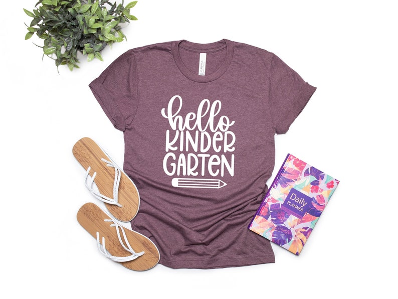 May include: A maroon t-shirt with the text "hello kindergarten" in white lettering and a white pencil graphic.