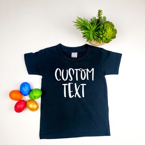 Puede incluir: Una camiseta negra con texto blanco que dice "CUSTOM TEXT". La camiseta est&aacute; rodeada de huevos de Pascua coloridos.