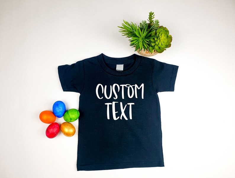 Puede incluir: Una camiseta negra con texto blanco que dice "CUSTOM TEXT". La camiseta est&aacute; rodeada de huevos de Pascua coloridos.