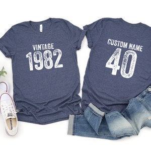 May include: Two blue t-shirts with white text. One shirt says "Vintage 1982" and the other says "Custom Name 40".