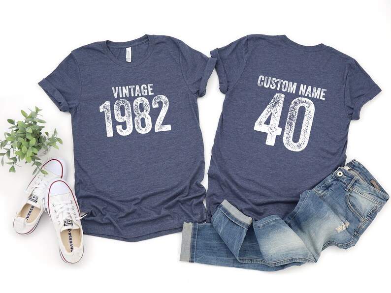 May include: Two blue t-shirts with white text. One shirt says "Vintage 1982" and the other says "Custom Name 40".