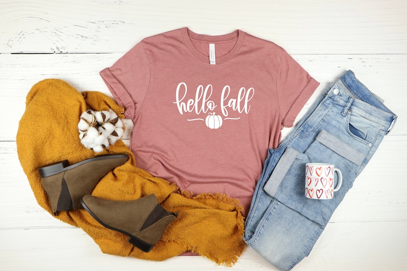 May include: A pink t-shirt with the text "hello fall" in white lettering and a white pumpkin graphic. The shirt is laid out on a white wooden surface with a pair of brown ankle boots, a yellow blanket, a cotton ball, a pair of blue jeans, and a white mug with red hearts.