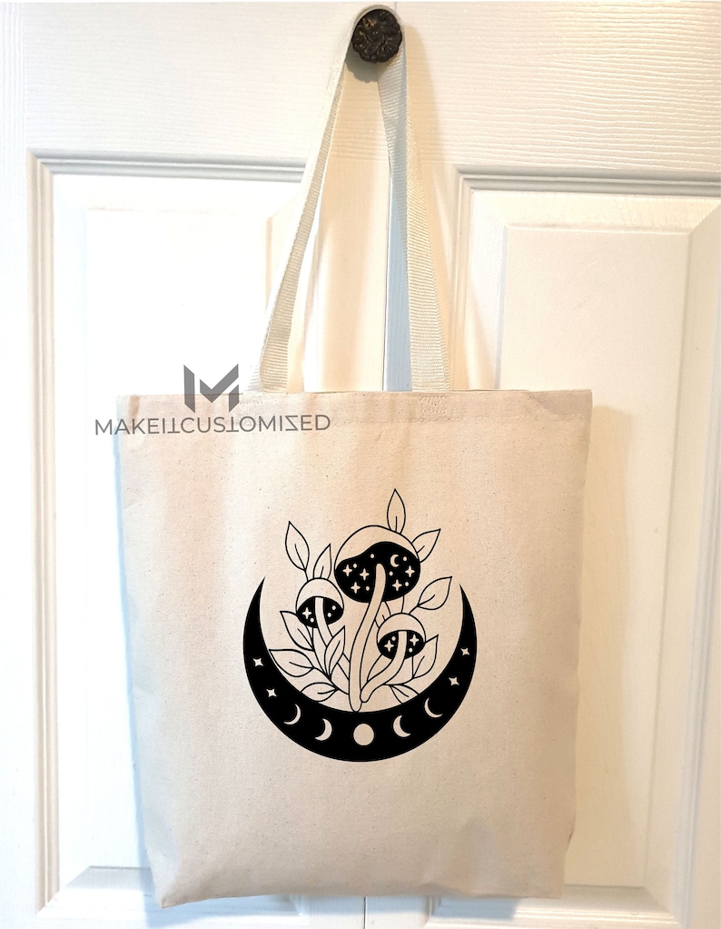 May include: A white canvas tote bag with black handles and a black graphic of mushrooms growing under a crescent moon with stars. The text "MAKEITCUSTOMIZED" is printed on the bag.
