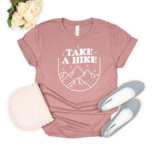 May include: A mauve t-shirt with the text "TAKE A HIKE" in white, featuring a stylized mountain range graphic inside a circle. The shirt is laid flat on a white background with a pair of striped canvas shoes.
