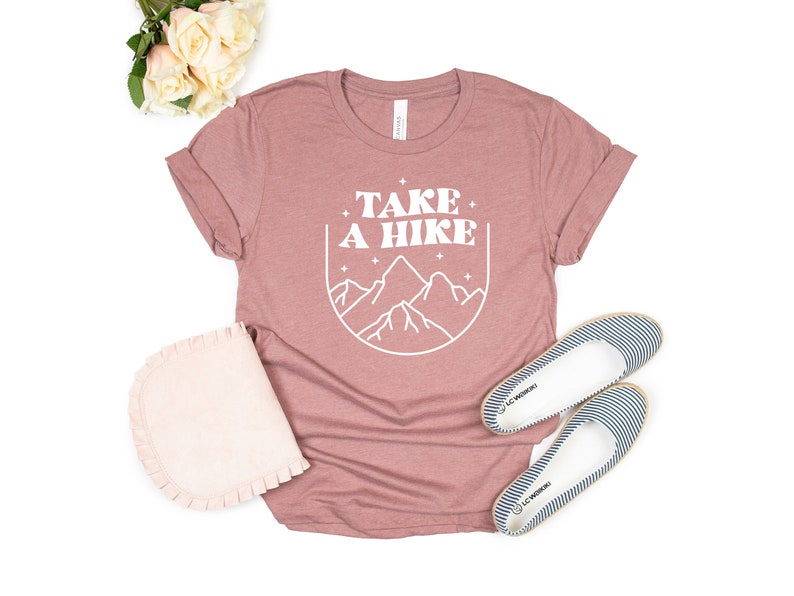 May include: A mauve t-shirt with the text "TAKE A HIKE" in white, featuring a stylized mountain range graphic inside a circle. The shirt is laid flat on a white background with a pair of striped canvas shoes.