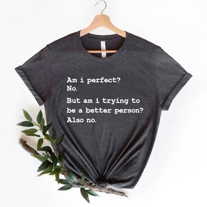 Puede incluir: Una camiseta gris oscuro con texto blanco que dice "&iquest;Soy perfecto? No. Pero, &iquest;estoy tratando de ser una mejor persona? Tampoco."