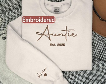 以下が含まれることがあります： 胸元にAuntie Est. 2025と茶色の刺繍が施されたライトベージュのクルーネックトレーナー。左袖にはoliviaという名前と小さなハートのシンボルが刺繍されています。衣類にはGildanのブランドラベルが付いています。