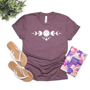May include: A maroon t-shirt with a white graphic of a moon phase design. The design features a crescent moon, a full moon, and a half moon.