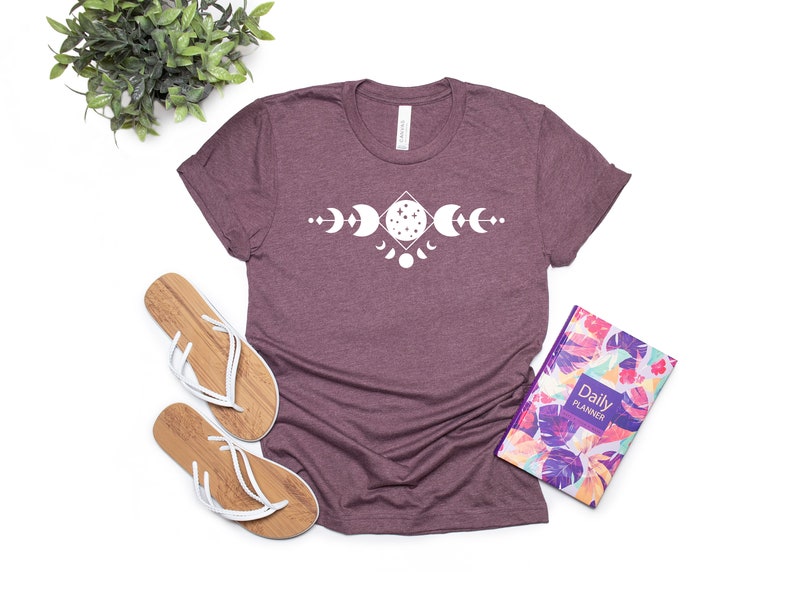 May include: A maroon t-shirt with a white graphic of a moon phase design. The design features a crescent moon, a full moon, and a half moon.