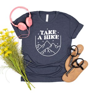 May include: A navy blue t-shirt with white text that says "TAKE A HIKE" and a graphic of mountains. The shirt is laying flat on a white background with a pair of sandals, a bouquet of yellow flowers, and pink headphones.