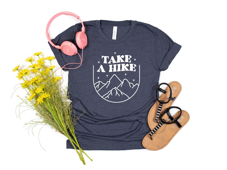 May include: A navy blue t-shirt with white text that says "TAKE A HIKE" and a graphic of mountains. The shirt is laying flat on a white background with a pair of sandals, a bouquet of yellow flowers, and pink headphones.