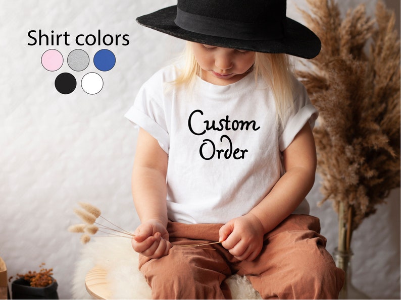 Puede incluir: Una camiseta blanca con el texto "Custom Order" en escritura negra. La camiseta la lleva puesta un ni&ntilde;o que lleva pantalones marrones y un sombrero negro. La imagen tambi&eacute;n muestra una tabla de colores con seis colores de camiseta diferentes: rosa, gris, azul, negro y blanco.