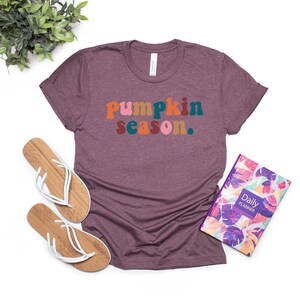 Może przedstawiać: Maroonowy t-shirt z napisem "pumpkin season." w kolorowych, retro literach.