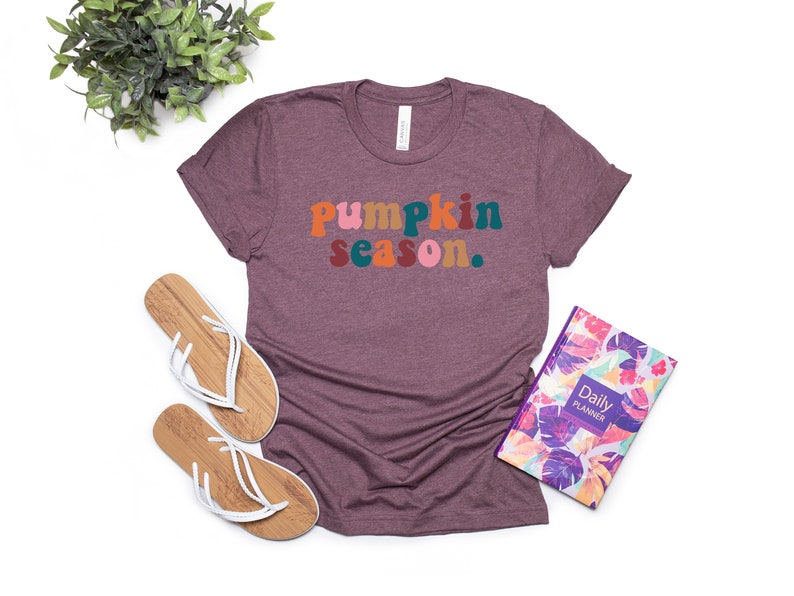 Może przedstawiać: Maroonowy t-shirt z napisem "pumpkin season." w kolorowych, retro literach.