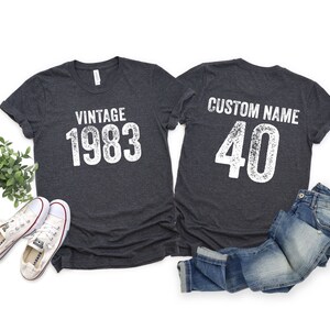 K&ouml;nnte beinhalten: Zwei dunkelgraue T-Shirts mit wei&szlig;em Text. Ein Shirt sagt "Vintage 1983" und das andere sagt "Benutzerdefinierter Name 40".