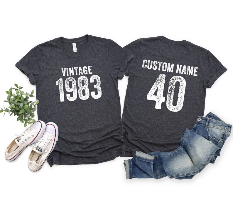 K&ouml;nnte beinhalten: Zwei dunkelgraue T-Shirts mit wei&szlig;em Text. Ein Shirt sagt "Vintage 1983" und das andere sagt "Benutzerdefinierter Name 40".