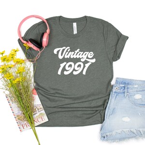 May include: A dark green t-shirt with the text "Vintage 1991" in white. The shirt is laid flat on a white background with a pair of pink headphones, a bouquet of yellow flowers, and a book.