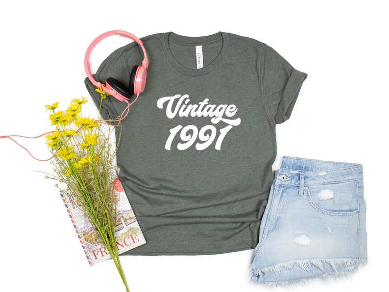 May include: A dark green t-shirt with the text "Vintage 1991" in white. The shirt is laid flat on a white background with a pair of pink headphones, a bouquet of yellow flowers, and a book.
