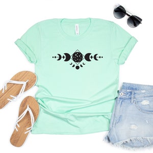 May include: A light green t-shirt with a black graphic of a moon and stars in a geometric design. The shirt is paired with denim shorts and white sandals.