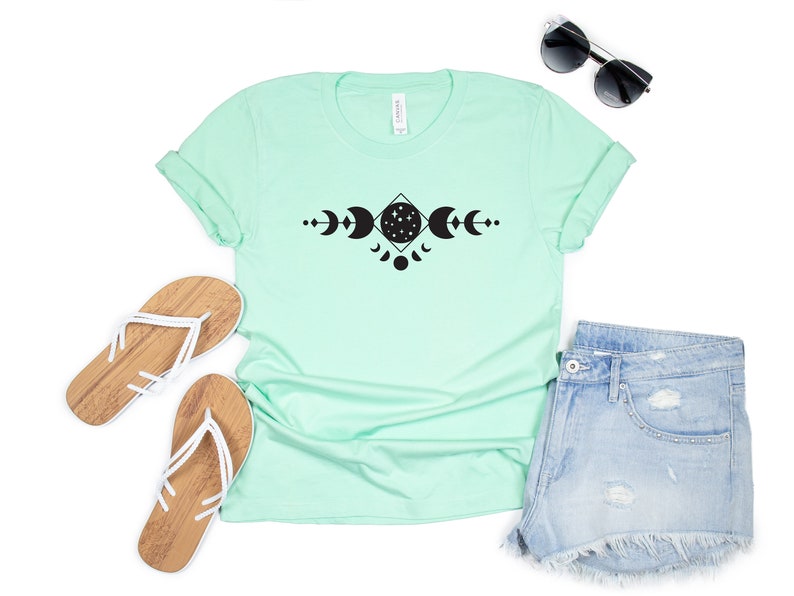 May include: A light green t-shirt with a black graphic of a moon and stars in a geometric design. The shirt is paired with denim shorts and white sandals.