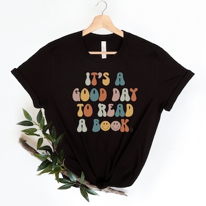 May include: Black t-shirt featuring a colourful retro-style graphic that reads "It's a good day to read a book." The graphic includes a smiley face with lightning bolts for eyes.