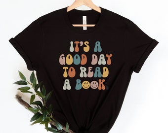 May include: Black t-shirt featuring a colourful retro-style graphic that reads "It's a good day to read a book." The graphic includes a smiley face with lightning bolts for eyes.