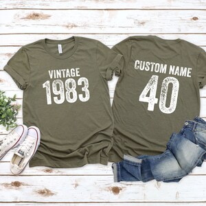 K&ouml;nnte beinhalten: Zwei olivgr&uuml;ne T-Shirts mit wei&szlig;em Text. Das vordere Shirt sagt "Vintage 1983". Das hintere Shirt sagt "Custom Name 40".