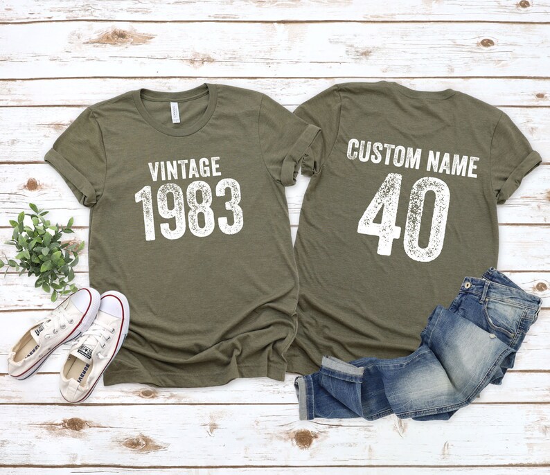 K&ouml;nnte beinhalten: Zwei olivgr&uuml;ne T-Shirts mit wei&szlig;em Text. Das vordere Shirt sagt "Vintage 1983". Das hintere Shirt sagt "Custom Name 40".