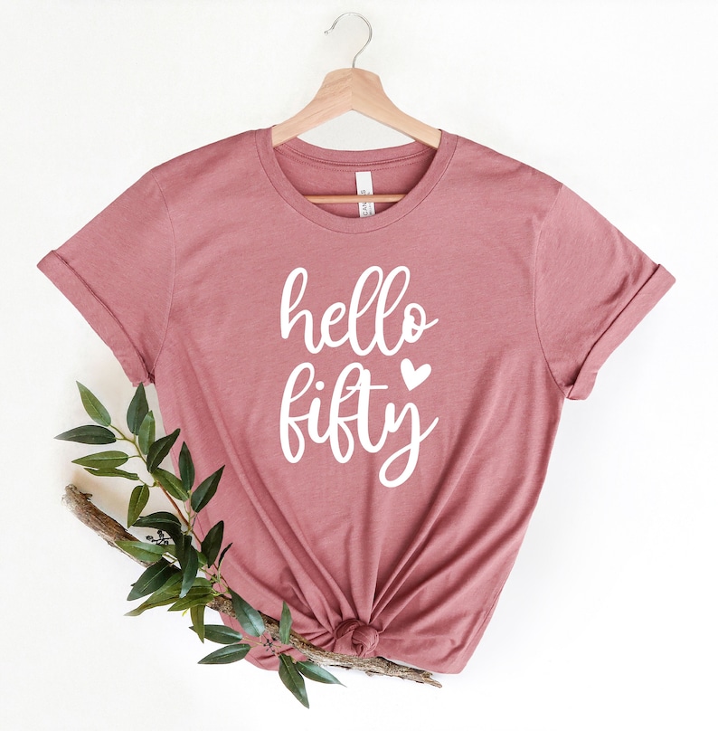 May include: A mauve t-shirt with the text "hello fifty" in white with a small heart next to the word "fifty".