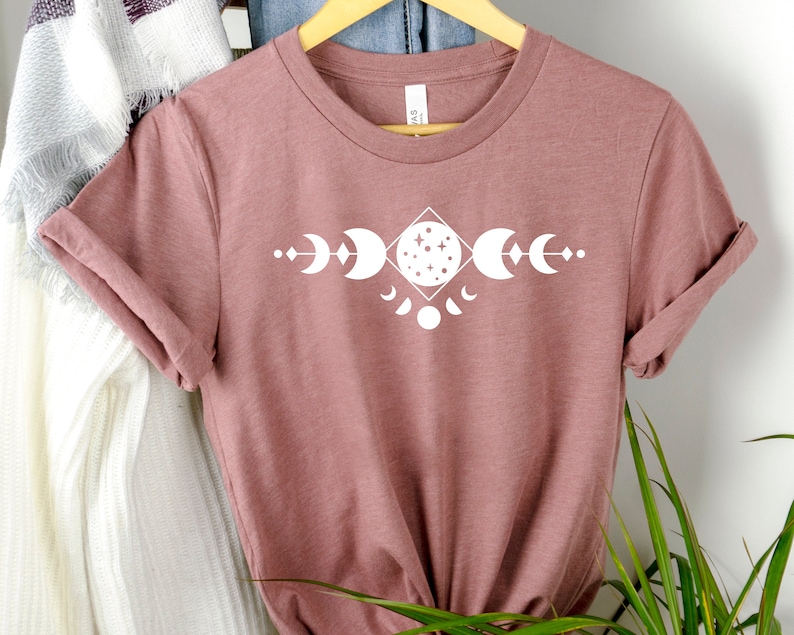 May include: A mauve colored t-shirt with a white graphic of a moon phase design. The design features a full moon with stars, crescent moons, and a half moon.