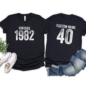 May include: Two black t-shirts with white text. The front shirt says "Vintage 1982". The back shirt says "Custom Name 40".