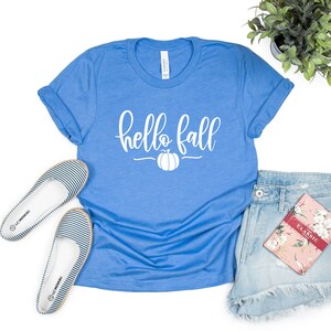 May include: A blue t-shirt with the text "hello fall" in white lettering and a white pumpkin graphic. The shirt is laid flat on a white background with a pair of striped flats, denim shorts, and a floral notebook.