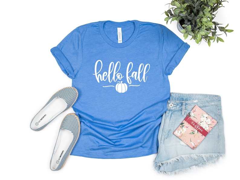May include: A blue t-shirt with the text "hello fall" in white lettering and a white pumpkin graphic. The shirt is laid flat on a white background with a pair of striped flats, denim shorts, and a floral notebook.