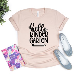 May include: A light pink t-shirt with the text "hello kindergarten" in black lettering and a black pencil graphic.