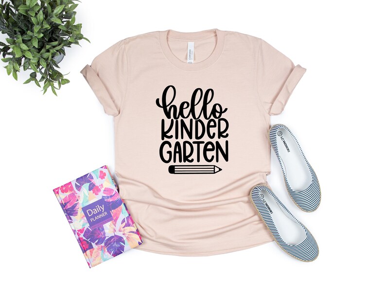 May include: A light pink t-shirt with the text "hello kindergarten" in black lettering and a black pencil graphic.