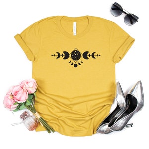 May include: A mustard yellow t-shirt with a black moon phases design. The design features a crescent moon, full moon, and a moon with stars inside. The moon phases are arranged in a geometric pattern.