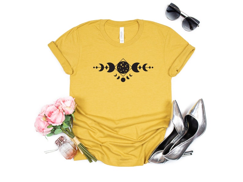May include: A mustard yellow t-shirt with a black moon phases design. The design features a crescent moon, full moon, and a moon with stars inside. The moon phases are arranged in a geometric pattern.